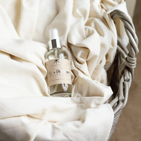 A spray bottle labeled "Fig and Pink Peppercorn Room and Linen Spray" from Flicker And Twinkle rests on soft, cream-colored fabric inside a woven basket. The luxury home fragrance, featuring a silver pump and a transparent body, suggests it contains a room and linen spray infused with the sophisticated scent of Fig and Pink Peppercorn. The scene conveys freshness and comfort.