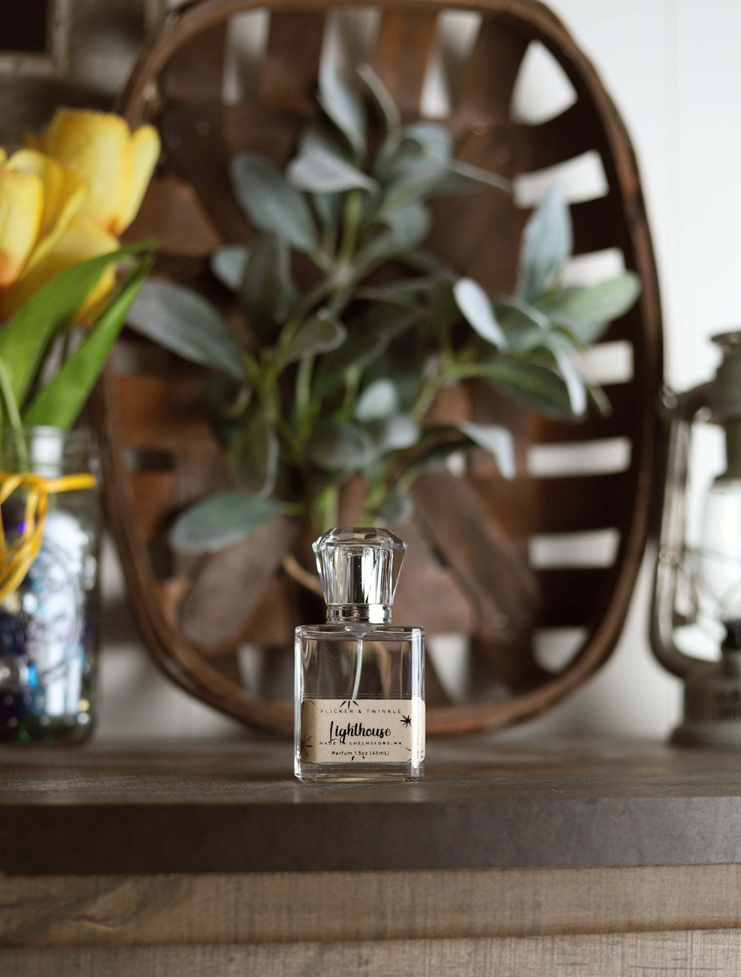 A glass bottle of Lighthouse Parfum by Flicker And Twinkle rests on a wooden surface, evoking the scent of ocean air. Behind it, a decorative wooden tray adorned with green leaves adds charm. To the left, a vase holds yellow tulips, and to the right, there's a metallic lantern.