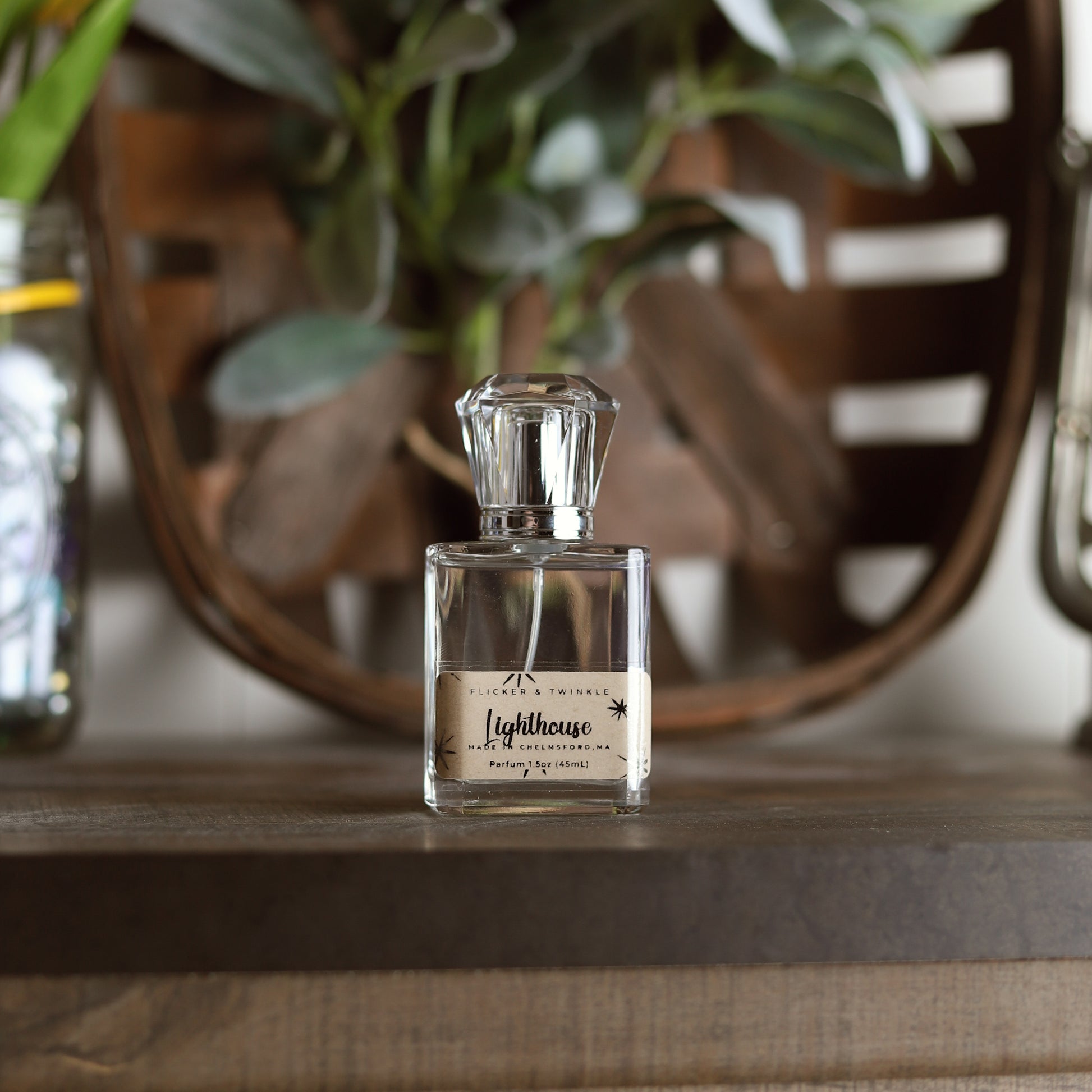 A perfume bottle labeled "Lighthouse Parfum" by Flicker And Twinkle rests on a wooden surface. The bottle features a clear, faceted cap, and the blurred green leaves in the background evoke hints of sea salt and ocean air.