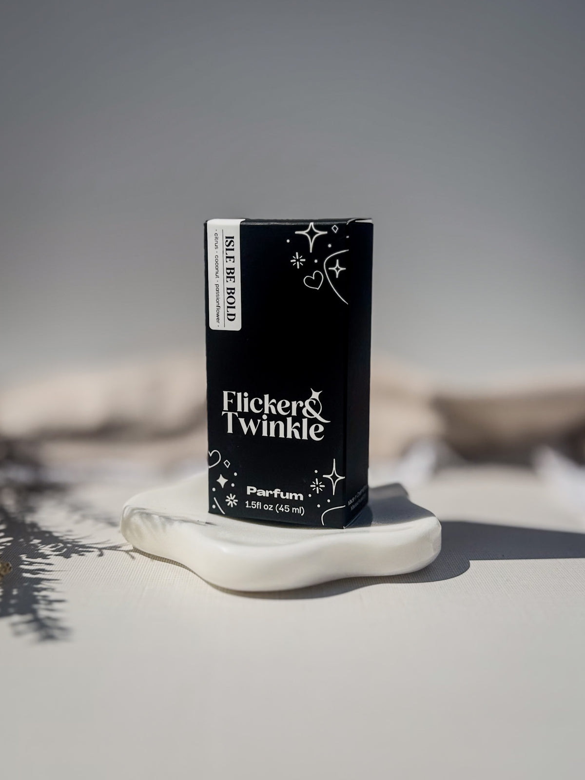 A black rectangular perfume box labeled 'Flicker Twinkle' and 'Isle Be Bold' stands upright on a white sculptural dish against a soft gray background. Isle be Bold is a passionflower and coconut scented parfum.