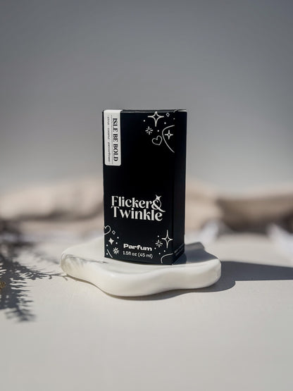 A black rectangular perfume box labeled 'Flicker Twinkle' and 'Isle Be Bold' stands upright on a white sculptural dish against a soft gray background. Isle be Bold is a passionflower and coconut scented parfum.