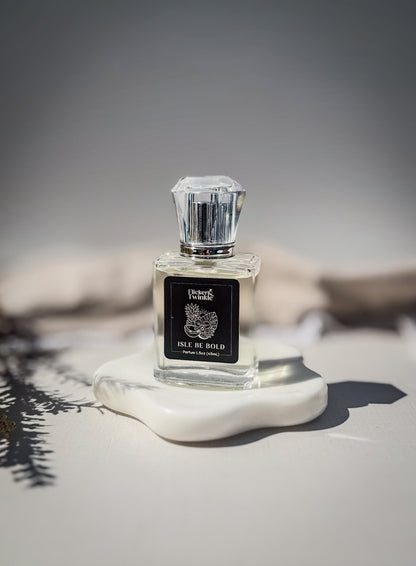 A bright tropical inspired Parfum fragrance called "Isle Be Bold" is in a clear glass perfume bottle with a black label on it. The perfume bottle is standing upright on a white ceramic dish with a soft beige and gray background. 