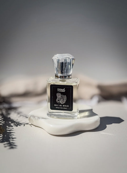 A bright tropical inspired Parfum fragrance called "Isle Be Bold" is in a clear glass perfume bottle with a black label on it. The perfume bottle is standing upright on a white ceramic dish with a soft beige and gray background. 