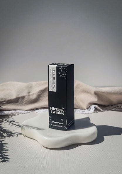 A black rectangular perfume box labeled 'Isle be Bold" stands on a white ceramic dish against a neutral gray background with draped fabric.