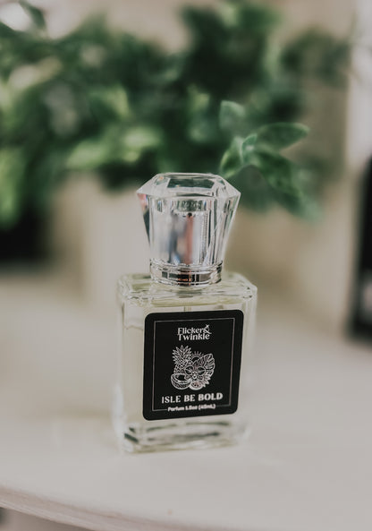 A clear glass perfume bottle with a faceted cap and a black label reading 'Isle Be Bold' Parfum, placed on a light surface with a blurred green plant in the background.