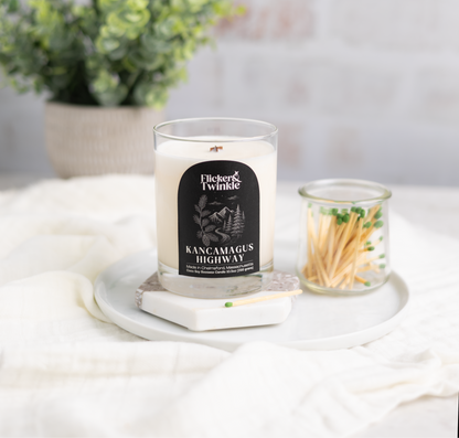 Kancamagus Highway Coconut Soy Beeswax Candle with a label on a coaster next to matches, with a plant in the background