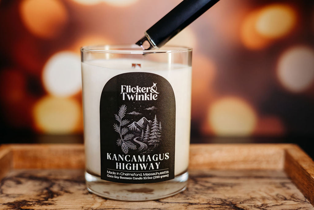 Coconut Soy Beeswax Candle labeled 'Flicker & Twinkle Kancamagus Highway' being lit on a wooden surface with a blurred background.