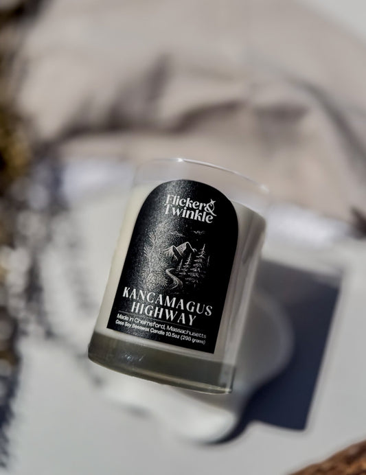 A clear glass jar candle with Coconut Soy Beeswax and a black label reading 'Flicker Twinkle' and 'Kancamagus Highway', photographed on a light surface with a blurred neutral background. Year round tree outdoor scent.
