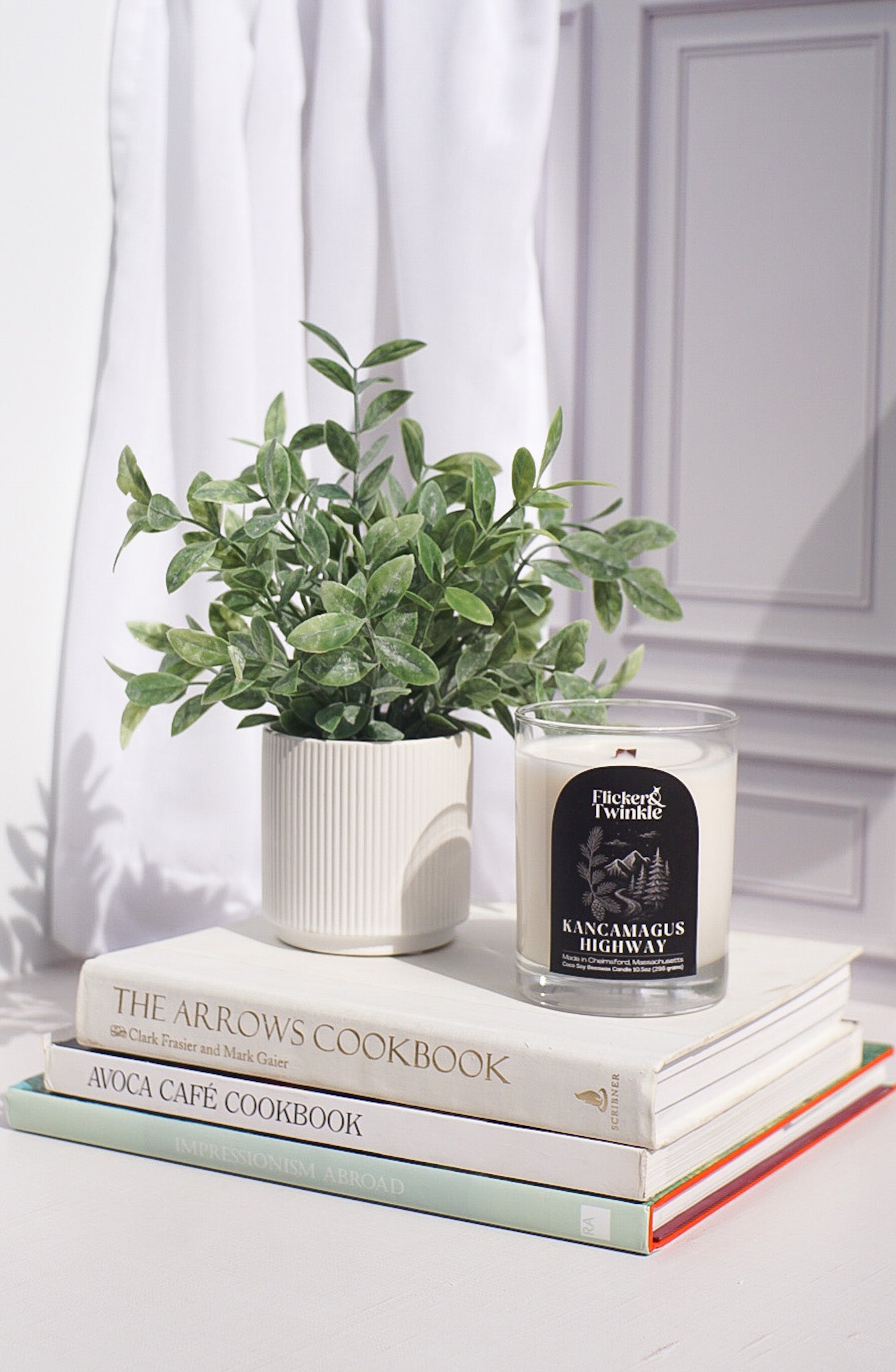 A potted plant and the Kancamagus Highway Candle atop cookbooks by a sunlit white curtain.