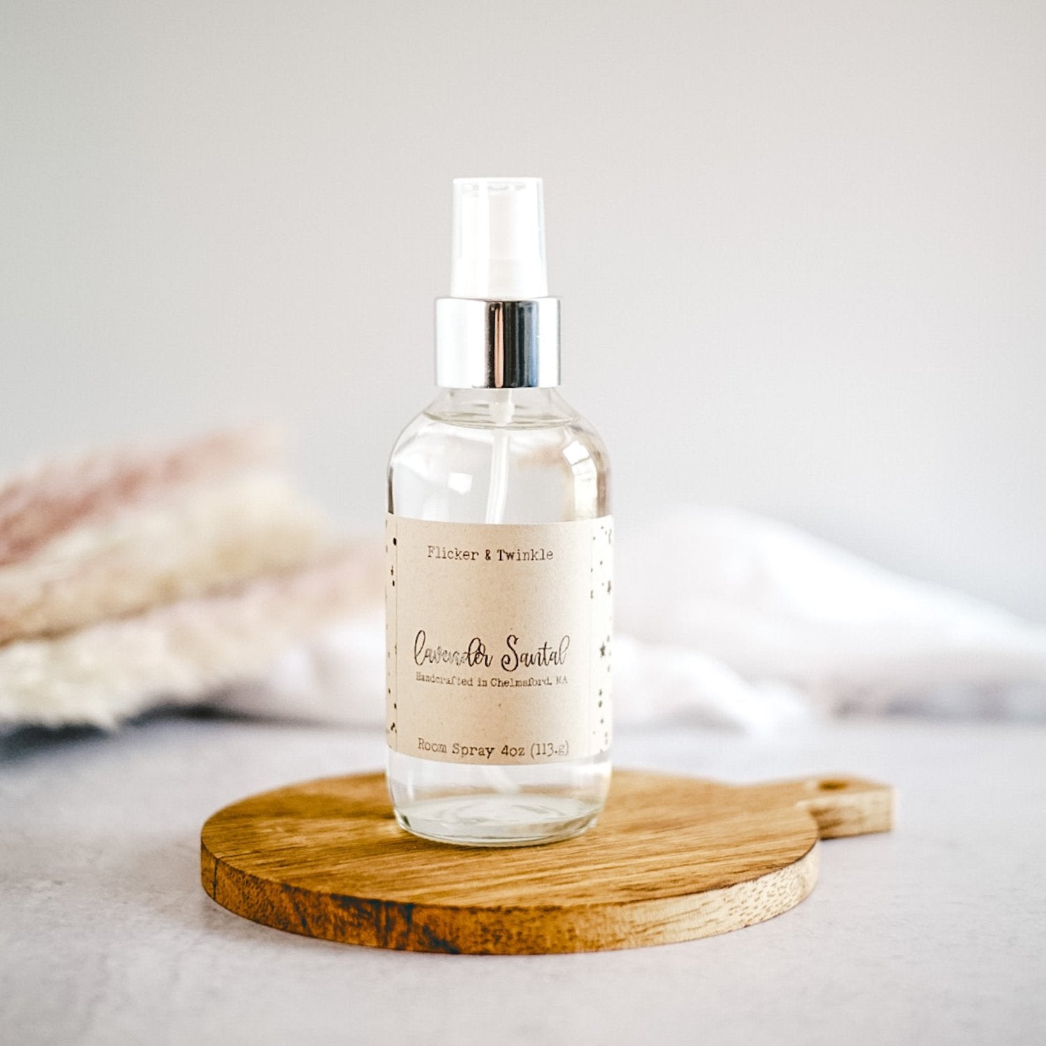 A clear spray bottle with a silver nozzle labeled "Lavender Santal Room Spray" from Flicker And Twinkle rests on a round wooden board. The backdrop is minimalistic, featuring soft, blurred elements that evoke a clean and serene ambiance. Each bottle is meticulously handpoured in small batches to ensure an artisanal, aromatherapeutic experience.