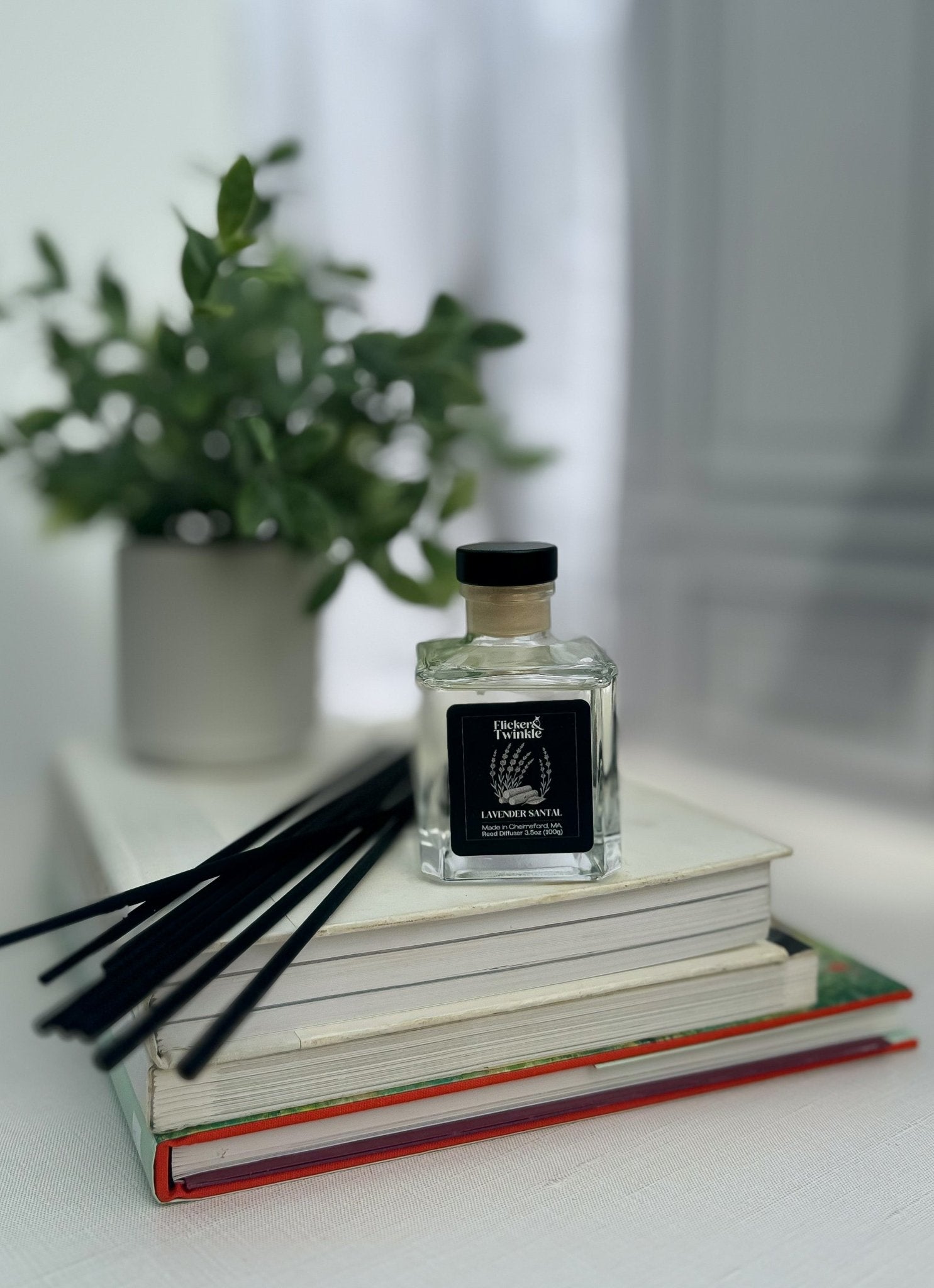 Lavender Santal Reed Diffuser with reeds sits on stacked books near a potted plant.