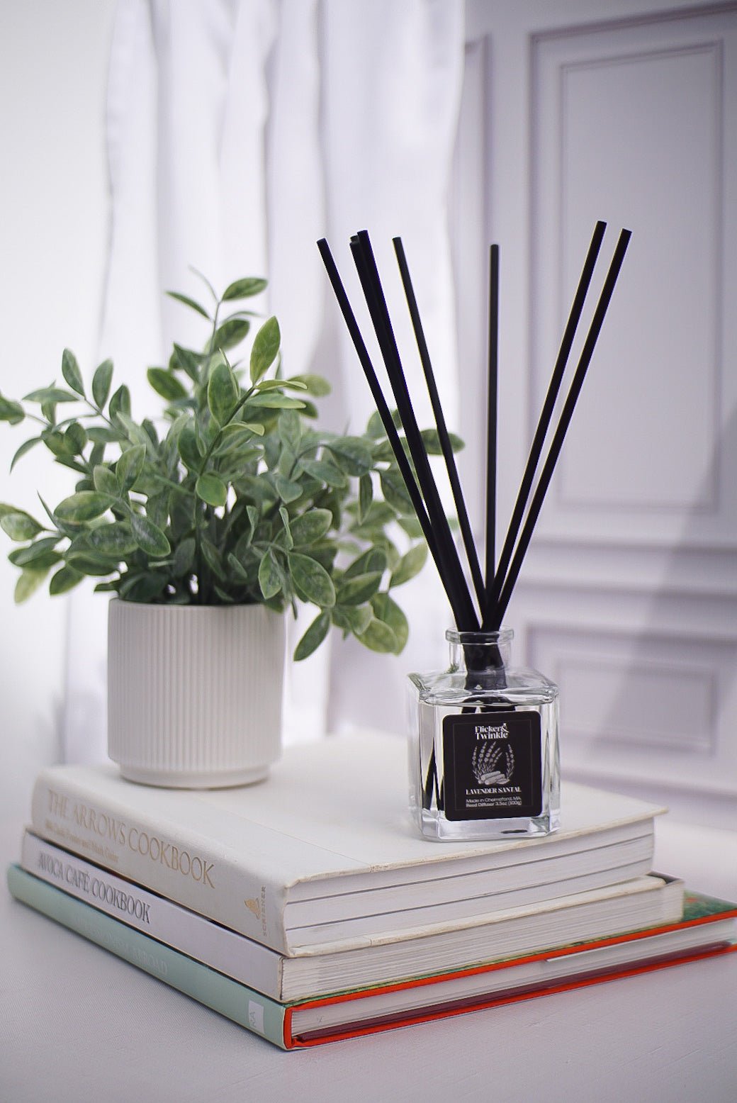 A potted plant and a Lavender Santal Reed Diffuser sit on three stacked cookbooks near a window.