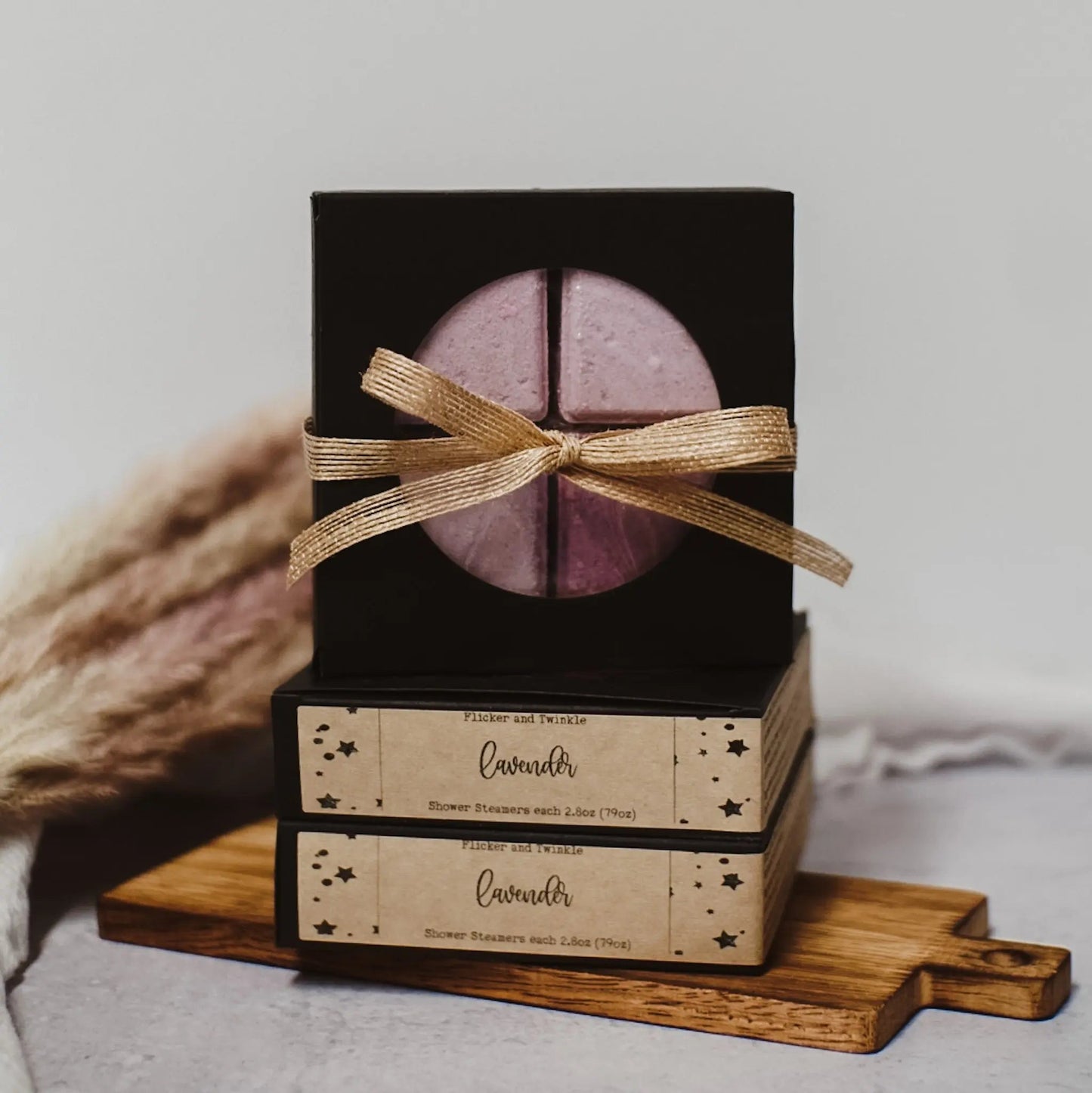 A neatly wrapped gift set with a bow containing Flicker And Twinkle's lavender-scented Shower Steamers is displayed on a small wooden cutting board. The set features a black box with a lavender steamer and two beige boxes with star patterns, all stacked for a spa-style escape.