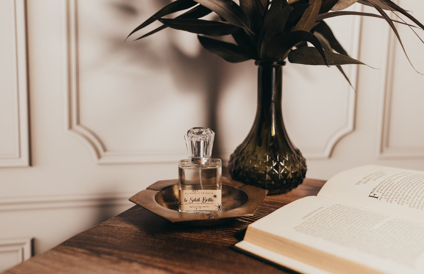 A bottle of Flicker And Twinkle's Le Soleil Brille Perfume sits on a small brass tray on a wooden table. Next to it is an open book, and in the background, there is a green vase with leaves. The setting exudes a cozy charm, with soft lighting and an elegant backdrop perfect for enjoying this exquisite summer fragrance.