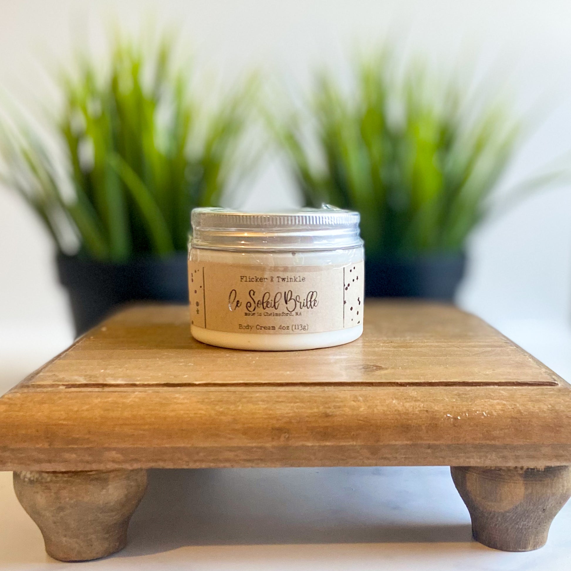 A jar of body cream labeled "Flicker and Twinkle Le Soleil Brille" sits on a small wooden platform, exuding a delicate summer fragrance. The background is blurred and features two potted plants with tall green leaves, casting a serene and natural ambiance.