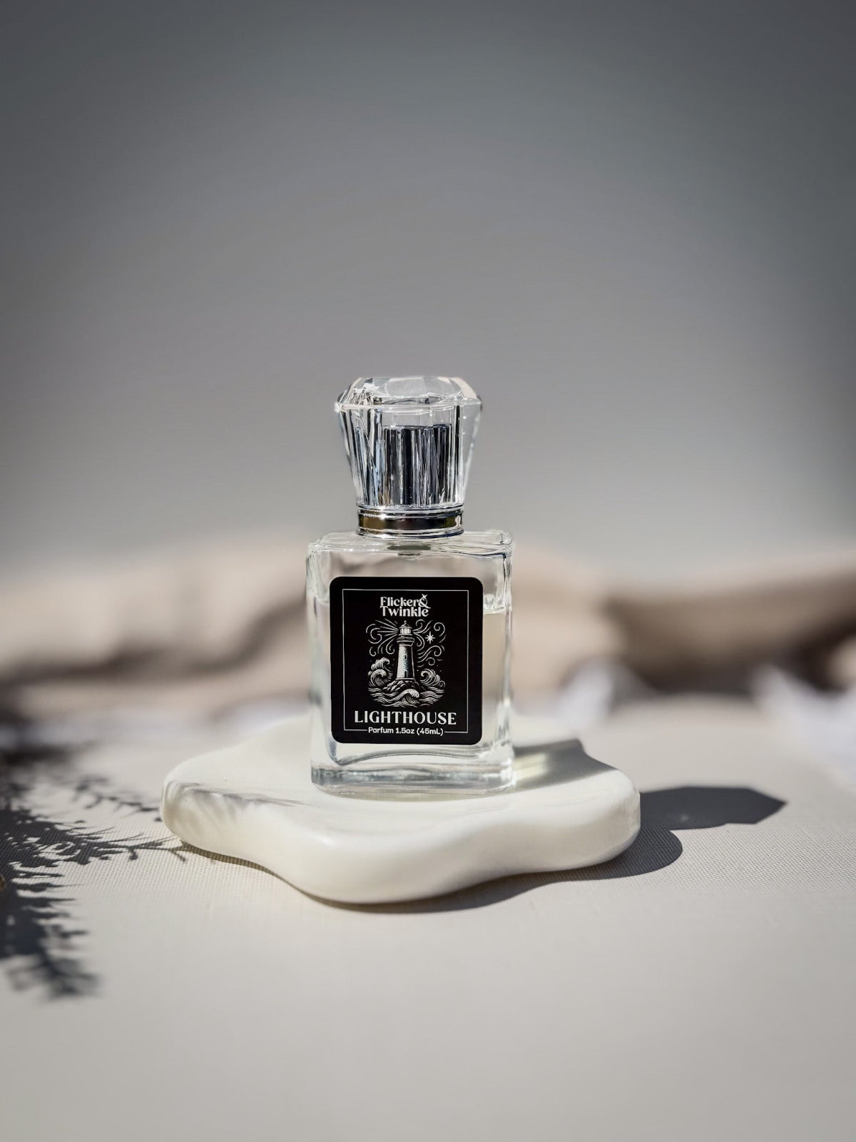 A clear glass perfume bottle with a faceted silver cap and a black label reading 'Lighthouse', displayed on a white stone slab against a soft gray background. This is a Clean white musk parfum fragrance.