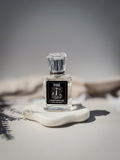 A clear glass perfume bottle with a faceted silver cap and a black label reading 'Lighthouse', displayed on a white stone slab against a soft gray background. This is a Clean white musk parfum fragrance.