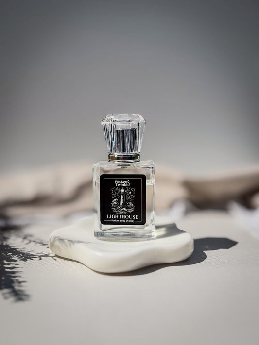 A clear glass perfume bottle with a faceted silver cap and a black label reading 'Lighthouse', displayed on a white stone slab against a soft gray background. This is a Clean white musk parfum fragrance.