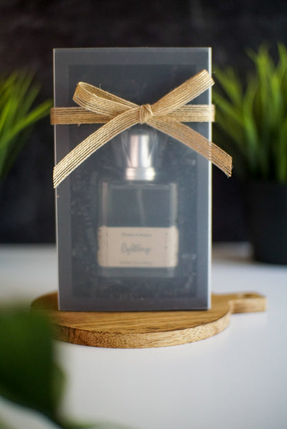 A Lighthouse Parfum bottle from Flicker and Twinkle is partially visible inside a transparent, plastic gift box wrapped with a gold ribbon tied in a bow. The gift box, evoking hints of sea salt and ocean air, is placed on a circular, wooden platform. In the background, there are green plants and a black wall.