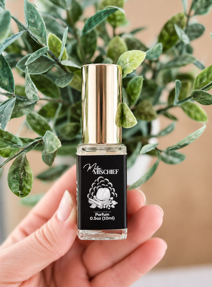 Mallow Mischief mini perfume has a black label with white writing and a picture of a marshmallow burning over a fire. This mini perfume is being held by a hand in front of a green plant. This scent smells like marshmallow matcha ginger perfume.