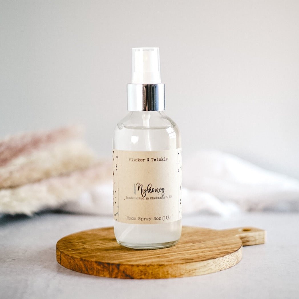 A clear bottle of Mykonos Room and Linen Spray by Flicker And Twinkle is placed on a wooden surface with a light-colored background. Infused with the Mykonos fragrance, this 4oz (113ml) bottle combines notes of citrus and fresh greens.