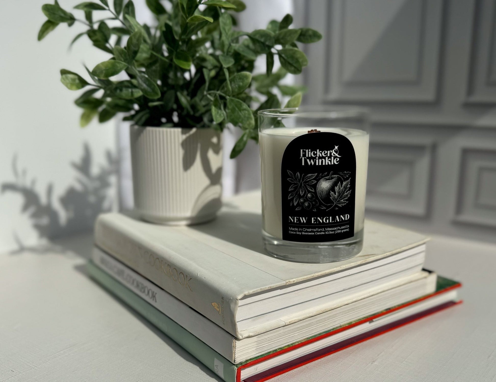 A potted plant and a New England Coconut Wax Candle sit atop books on a white surface.