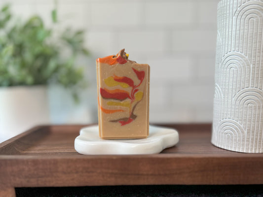 A rectangular bar of Flicker And Twinkle's New England Goat Milk Soap, inspired by the vibrant hues of a New England autumn and adorned with swirls of red, orange, yellow, and brown, stands upright on a minimalist white soap dish with rounded edges. In the background, soft focus greenery and a textured white cylindrical container enhance the scene.