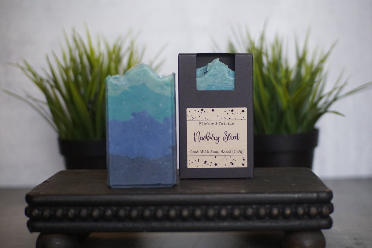 A handcrafted bar of Newbury Street Goat Milk Soap by Flicker And Twinkle showcases a gradient of blue hues, partially unboxed beside its minimalist black packaging labeled "Newbury Street Goat Milk Soap." This elegant display stands on a decorative surface, with green plants blurred in the background—perfect for the luxury connoisseur.