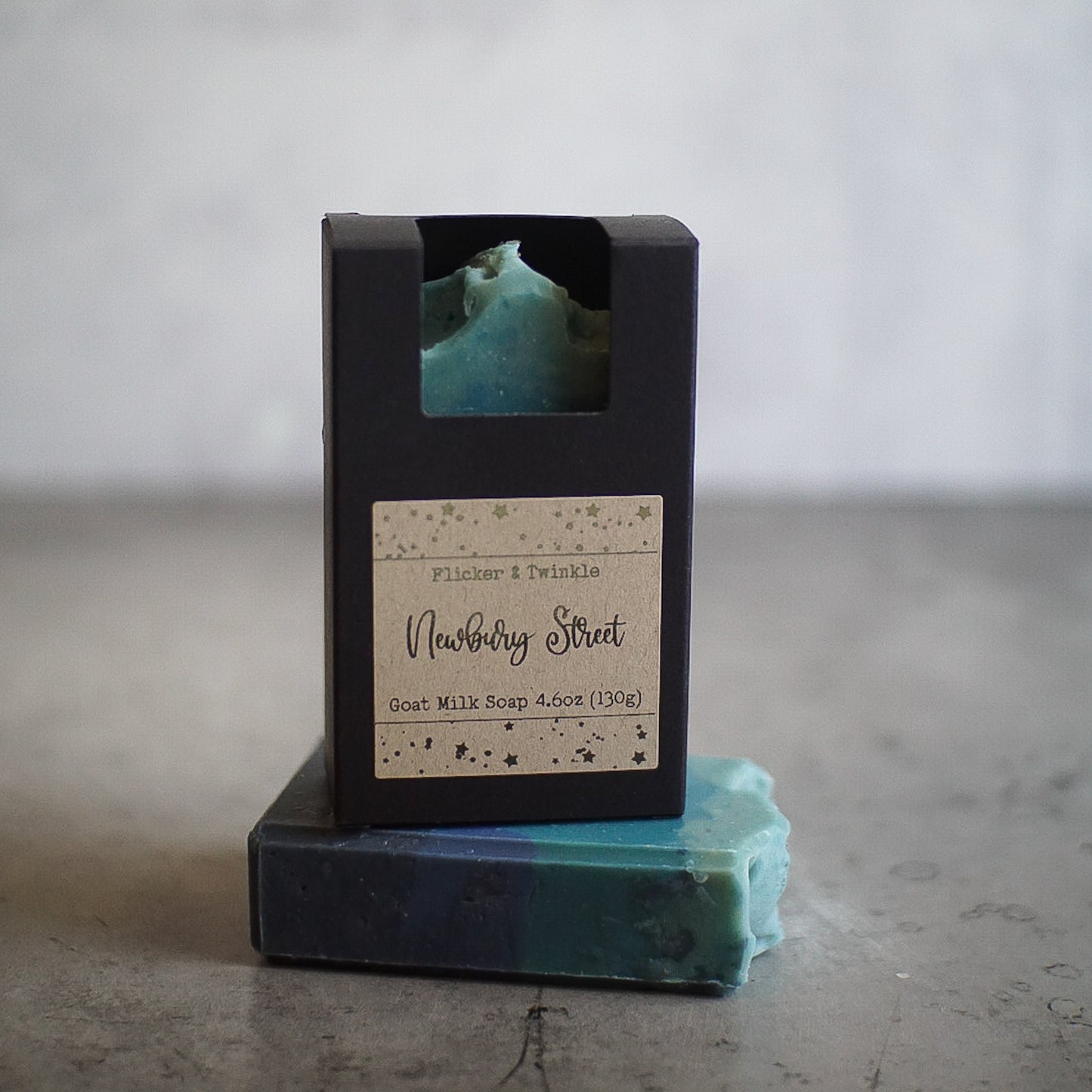 A bar of Newbury Street Goat Milk Soap sits in an open black box labeled "Flicker & Twinkle, Newbury Street, Goat Milk Soap, 4.6oz (130g)." Another similar teal and blue marbled soap bar lies flat on a textured gray surface next to the boxed soap—an ideal choice for the luxury connoisseur.