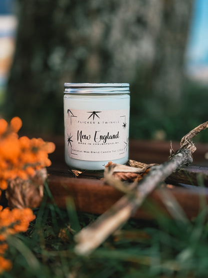 A jar labeled "New England Coconut Wax Candle," made by Flicker And Twinkle, sits on grass near an orange flower and a tree trunk. The jar is decorated with star drawings and has a silver lid. A twig lies beside the candle, evoking the essence of a crisp apple orchard.