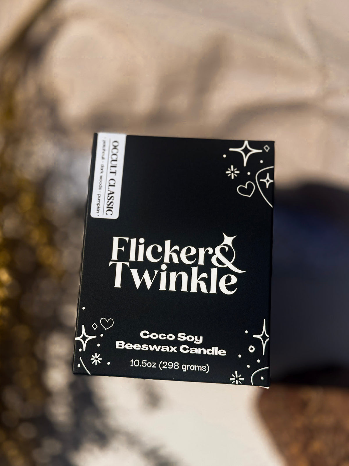 A black box of Flicker & Twinkle Coco Soy Beeswax Candle, 10.5 oz, shown against a blurred beige and brown background.