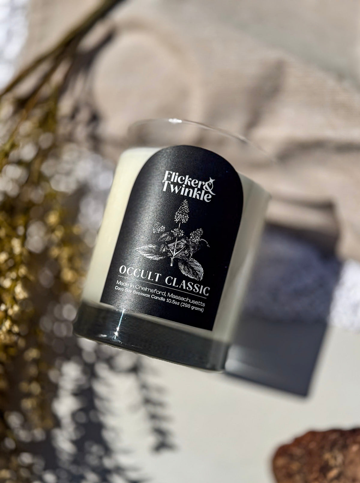 A Flicker & Twinkle 'Occult Classic' soy candle in a clear glass jar with a black label, styled on a tabletop with dried botanicals and neutral fabric in the background.
