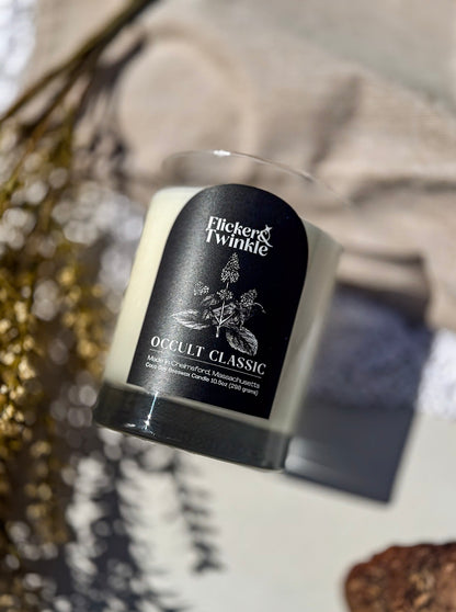 A Flicker & Twinkle 'Occult Classic' soy candle in a clear glass jar with a black label, styled on a tabletop with dried botanicals and neutral fabric in the background.