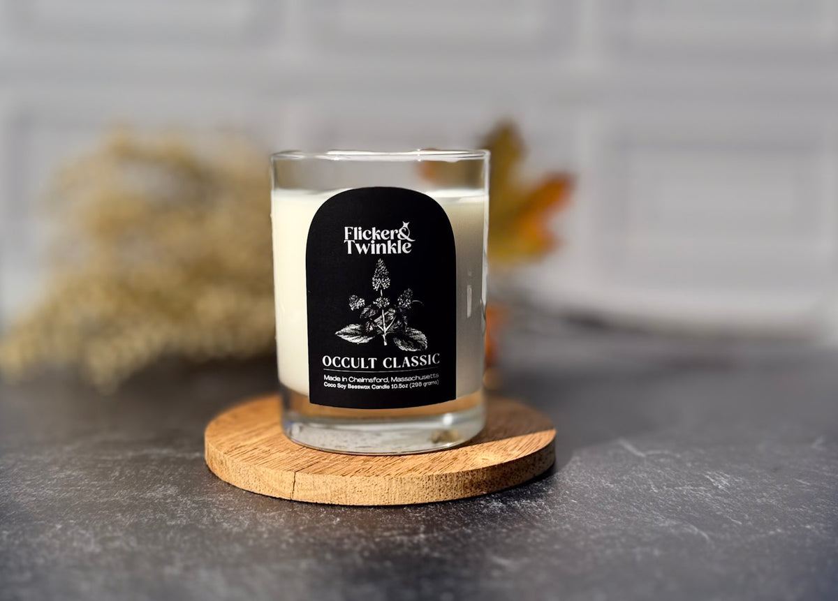 A glass jar candle with white wax sits on a round wooden coaster. The candle has a black label reading 'Flicker & Twinkle Occult Classic.