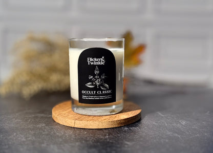 A glass jar candle with white wax sits on a round wooden coaster. The candle has a black label reading 'Flicker & Twinkle Occult Classic.