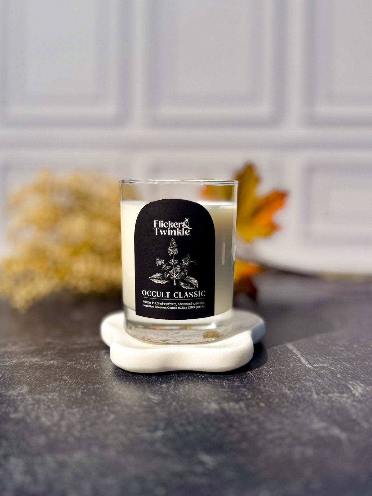 A Flicker & Twinkle 'Occult Classic' soy candle in a clear glass jar, resting on a white ceramic coaster on a dark gray stone surface with a blurred white paneled wall and autumn leaves in the background.