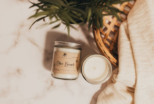 A cozy scene features two small batch handmade candles labeled "Olive Branch Coconut Wax Candle" by Flicker and Twinkle on a marble countertop. One candle is in a glass jar with a lid, while the other is an open candle without a lid. Nearby, a beige cloth and part of a green plant enhance the earthy and fruity scents.