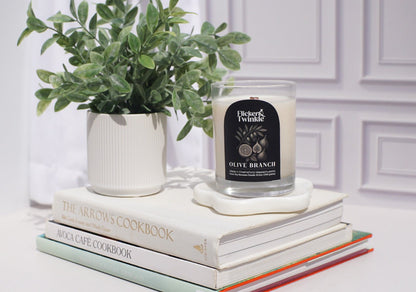 A potted plant and an Olive Branch Coconut Wax Candle sit on stacked cookbooks by the wall.