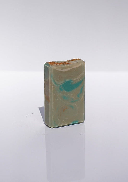 The Olive Branch Goat Milk Soap bar with blue and beige swirls stands upright on a white surface.