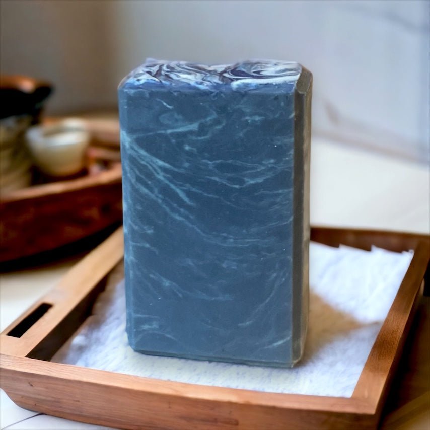 A rectangular block of The Yachtsman Goat Milk Soap by Flicker And Twinkle, featuring marbled black with white swirls, is displayed on a wooden tray with a white towel underneath. The background includes a blurred view of small bowls and a wooden container, creating an inviting scene reminiscent of an oceanic scent.