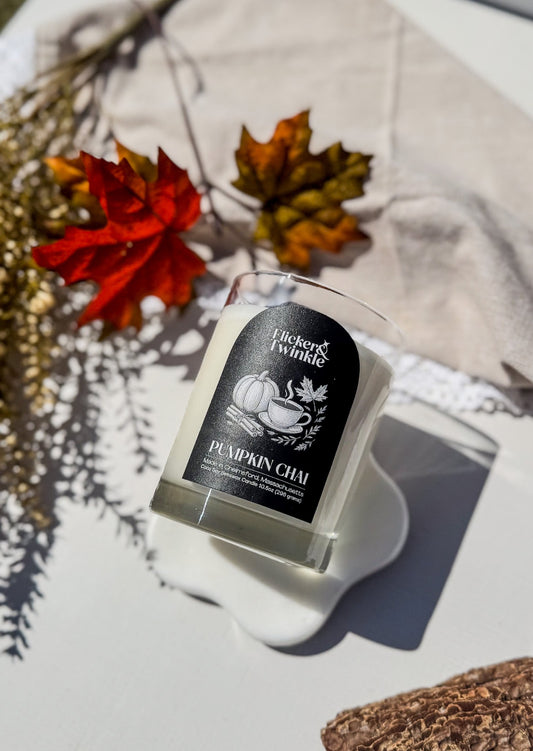 A glass jar candle made by Flicker and Twinkle scented in Pumpkin Chai. The Coconut soy wax candle sits on a white surface with autumn leaves, a beige cloth in a flat lay image.