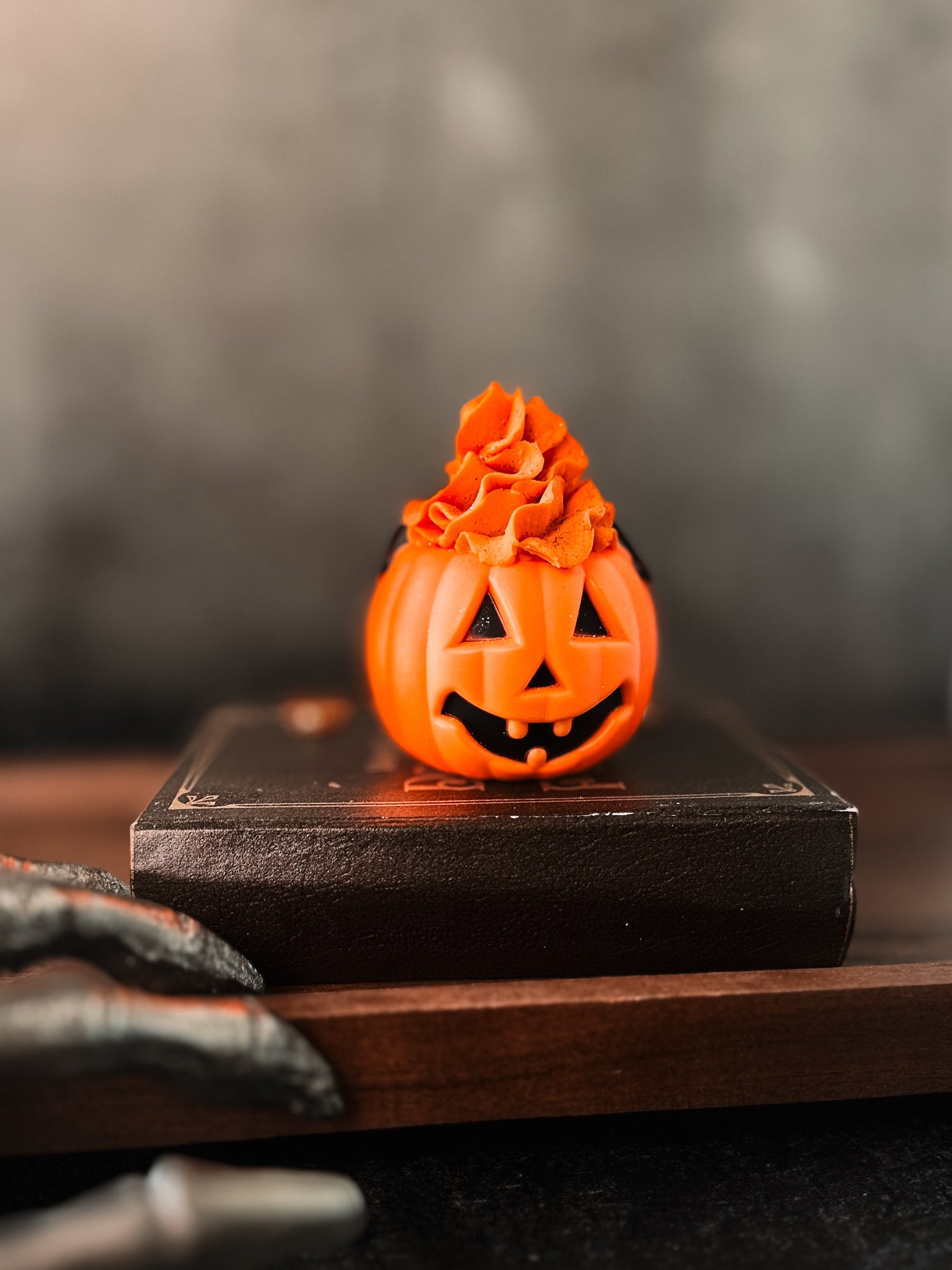 A small pumpkin-themed cupcake with orange frosting on top sits on a closed book. The cupcake has a jack-o'-lantern face with a happy expression. In the foreground, part of a claw-like decoration is visible, evoking vibes reminiscent of Flicker And Twinkle's Pumpkin Bath Bomb Cauldron, while the background is blurred.