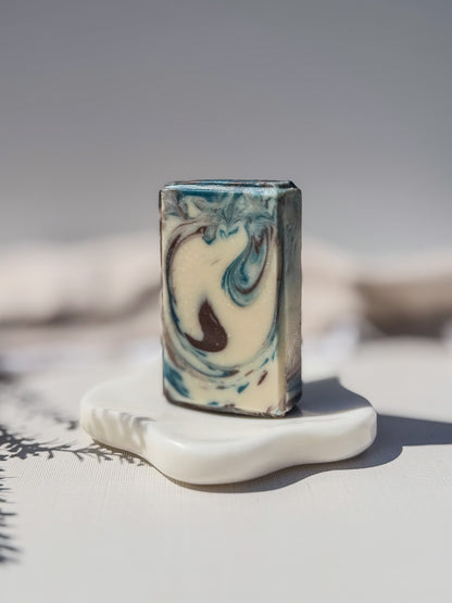 A rectangular artisan soap bar with a marbled swirl pattern of cream, blue, and dark brown, displayed on a small white ceramic stand.
