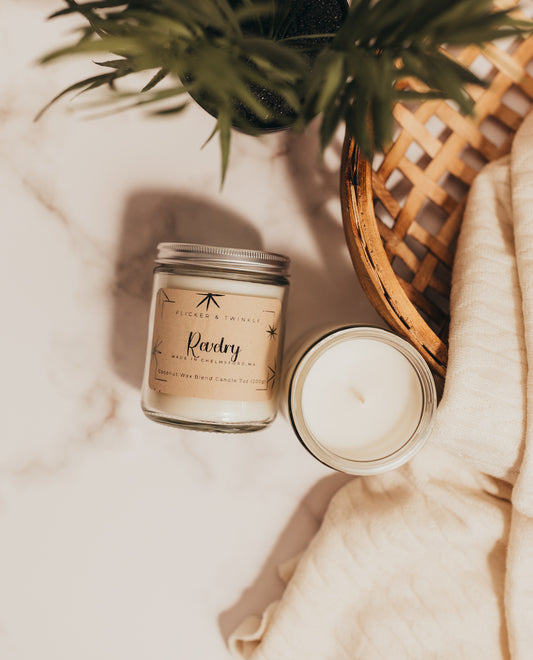 A Revelry Coconut Wax Candle by Flicker And Twinkle in a glass jar with a lid sits on a marble surface next to an open candle. A knit blanket, potted plant, and wicker tray are partially visible, adding a cozy feel to the scene. The label on the closed candle features a minimalist design mentioning sandalwood.