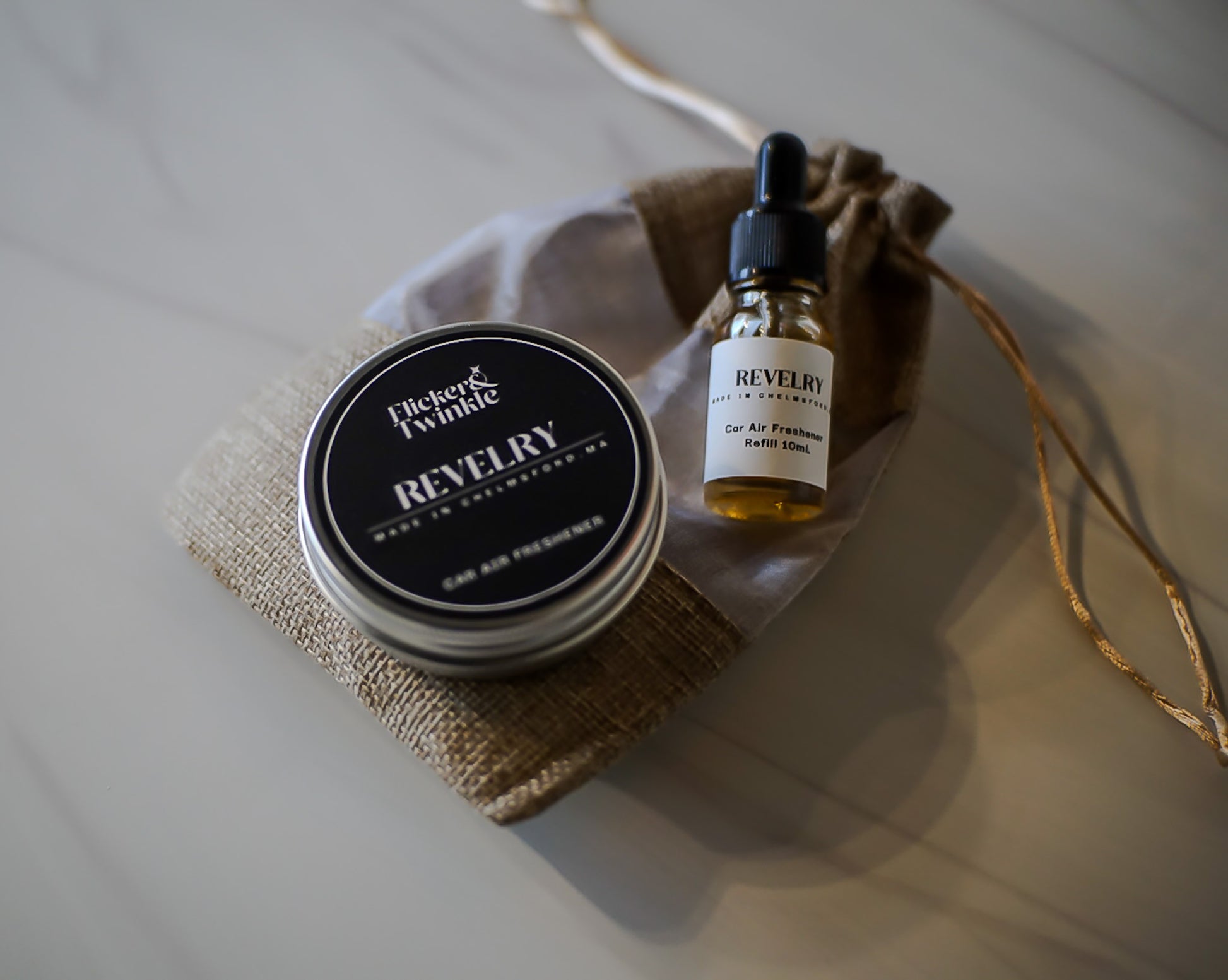 Car Freshenr tin and dropper bottle with 'Revelry' scent on a textured surface