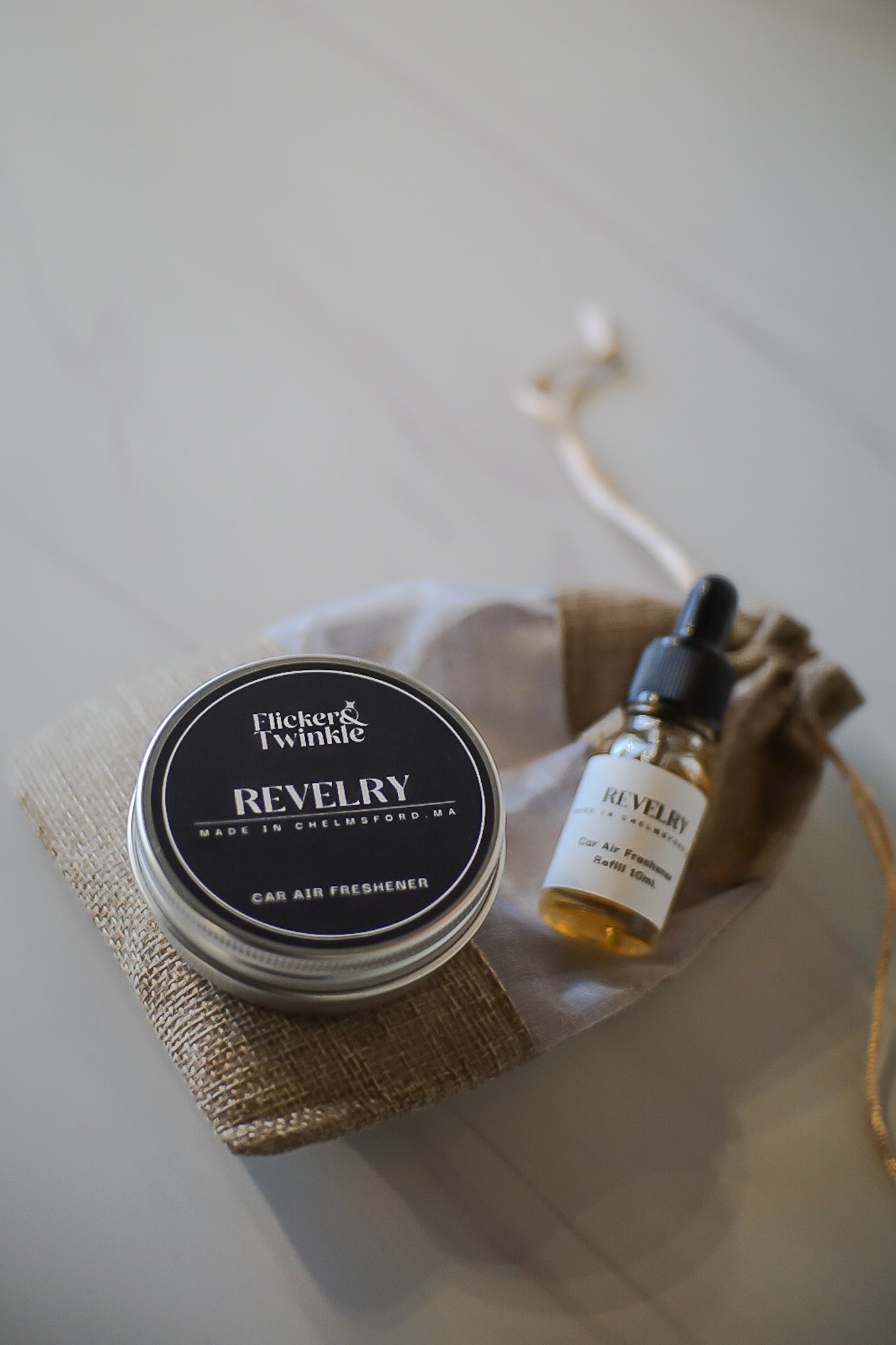 Two products, a small jar and a dropper bottle, on a white surface with a neutral background. Revelry Car freshener and refill for a long lasting Car Scent
