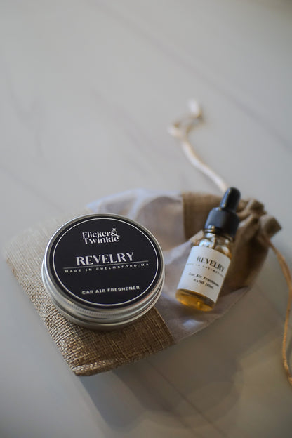 Two products, a small jar and a dropper bottle, on a white surface with a neutral background. Revelry Car freshener and refill for a long lasting Car Scent