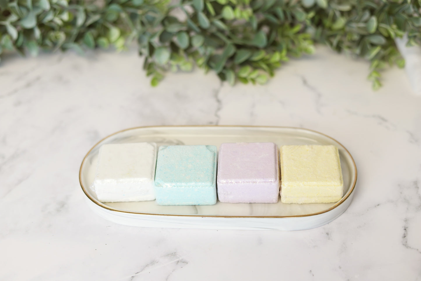 A white rectangular plate placed on a marble surface holds four pastel-colored square shower steamers by Flicker And Twinkle, infused with essential oils, arranged in a row. The colors are white, light blue, lavender, and cream. Greenery background decorates the scene.