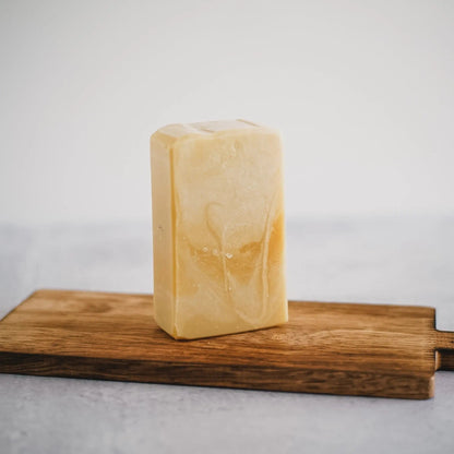 A rectangular, beige bar of "From Sorrento with Love" Goat's Milk Soap by Flicker and Twinkle, featuring subtle marbling, stands upright on a wooden cutting board. The background is simple and plain, focusing attention on the soap and the board.