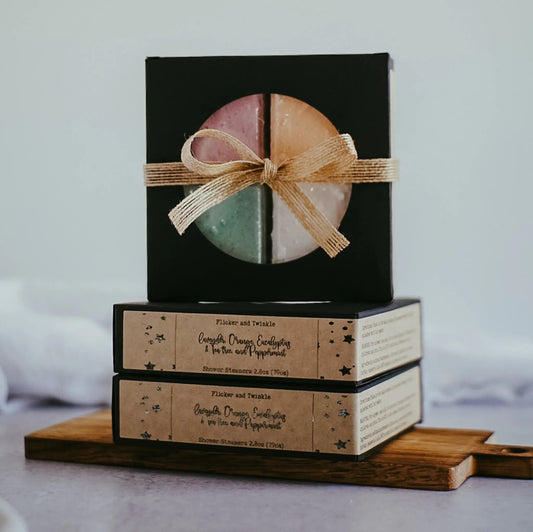 A stack of three square, black boxes from Flicker And Twinkle, with the top box tied with a ribbon, holds colorful circular Shower Steamers infused with essential oils. The lower two boxes feature beige labels decorated with stars and text. This elegant arrangement is presented on a wooden board against a neutral background, inviting you to a spa-style escape.